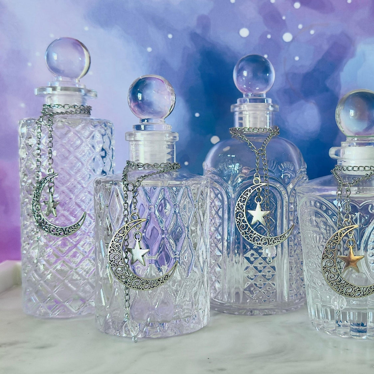 Five clear glass apothecary bottles with spherical stoppers and silver crescent moon and star chain ornaments, arranged on a light surface against a purple and blue starry background.