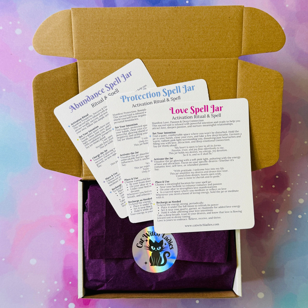An open kraft cardboard box containing purple tissue paper, a round branded sticker, and three printed spell jar cards labeled Abundance Spell Jar, Protection Spell Jar, and Love Spell Jar, set against a pastel galaxy background.