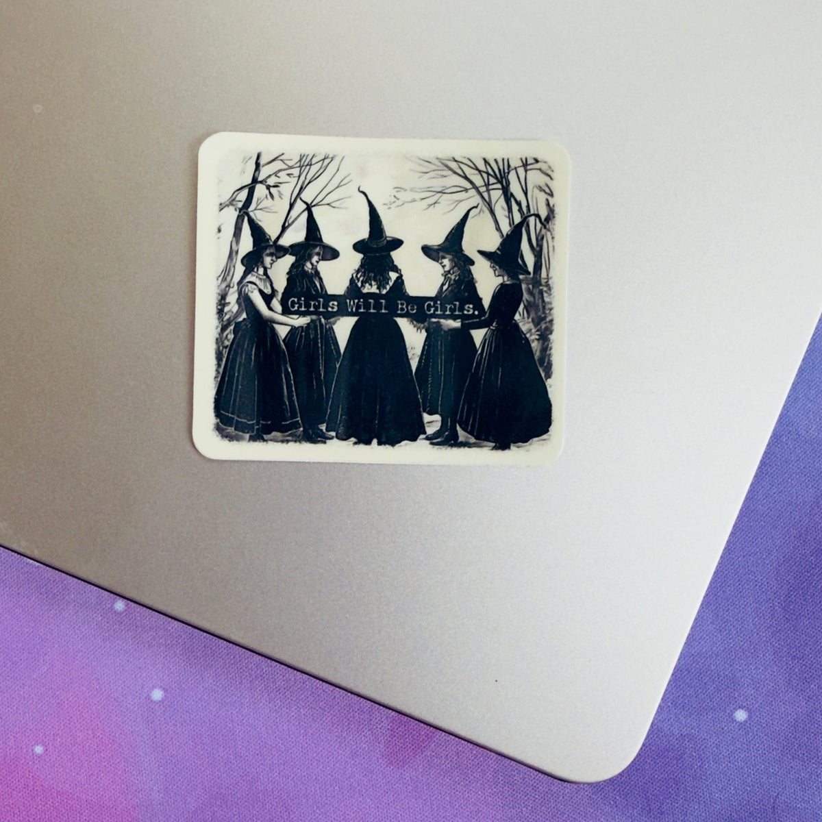 A rectangular sticker with rounded corners displayed on the corner of a silver laptop. The sticker features a black and white illustration of five witches in pointed hats standing together with bare trees in the background and the text 'Witches Will Be Visited'.