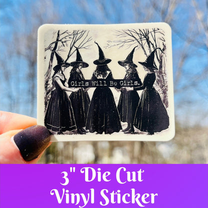 A hand holds a square die-cut vinyl sticker featuring a black-and-white illustration of five witches in pointed hats with the text 'Girls Will Be Girls.' The background is an outdoor scene with blurred trees and blue sky, and a purple banner at the bottom reads '3" Die Cut Vinyl Sticker'.