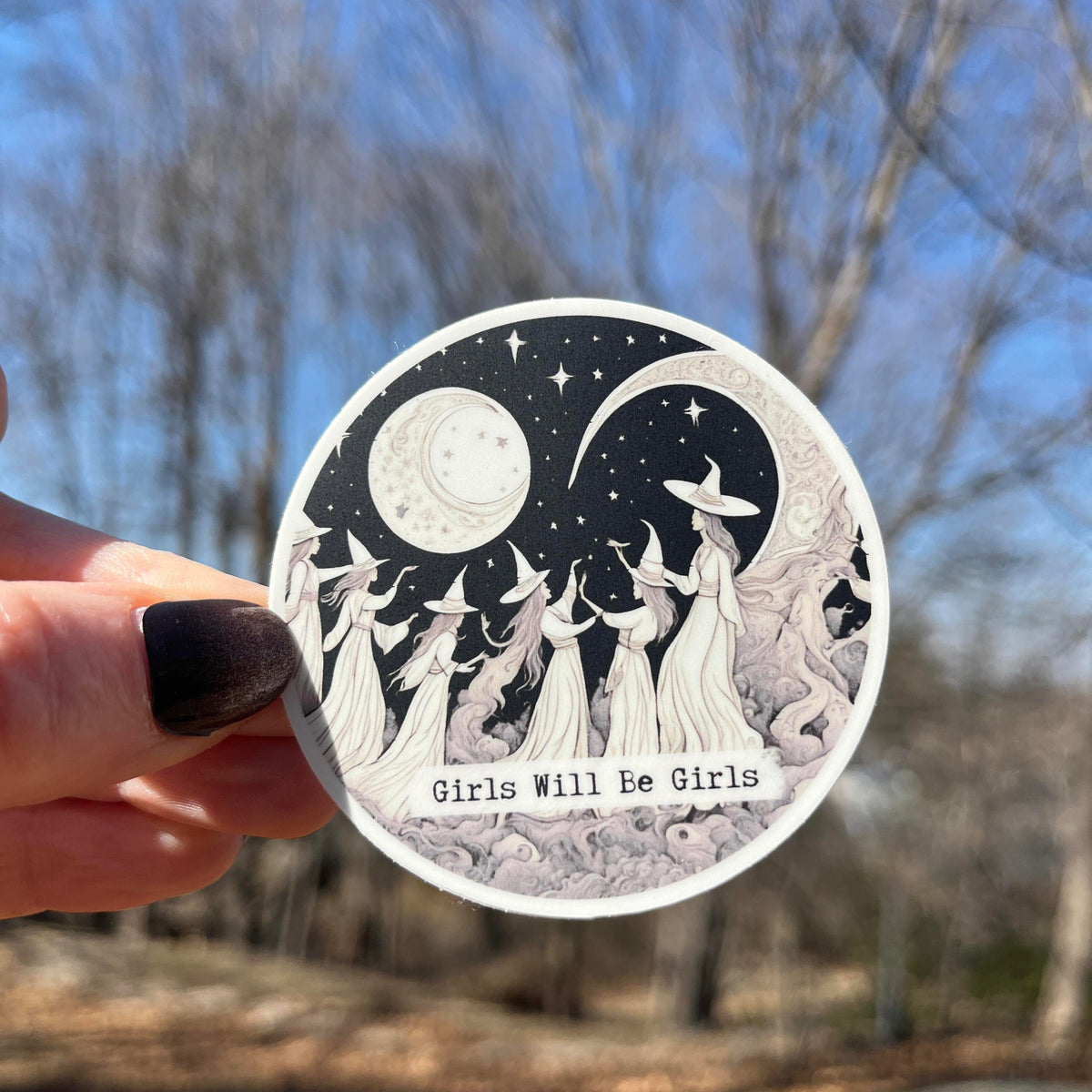 A hand with dark nail polish holds a round sticker featuring a black and white illustration of girls in witch hats under a crescent moon and stars, with the text 'Girls Will Be Girls' against a blurred outdoor background of trees and blue sky.