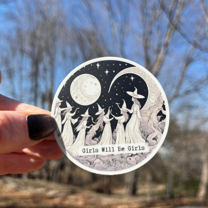 A hand with dark nail polish holds a round sticker featuring a black and white illustration of girls in witch hats under a crescent moon and stars, with the text 'Girls Will Be Girls' against a blurred outdoor background of trees and blue sky.