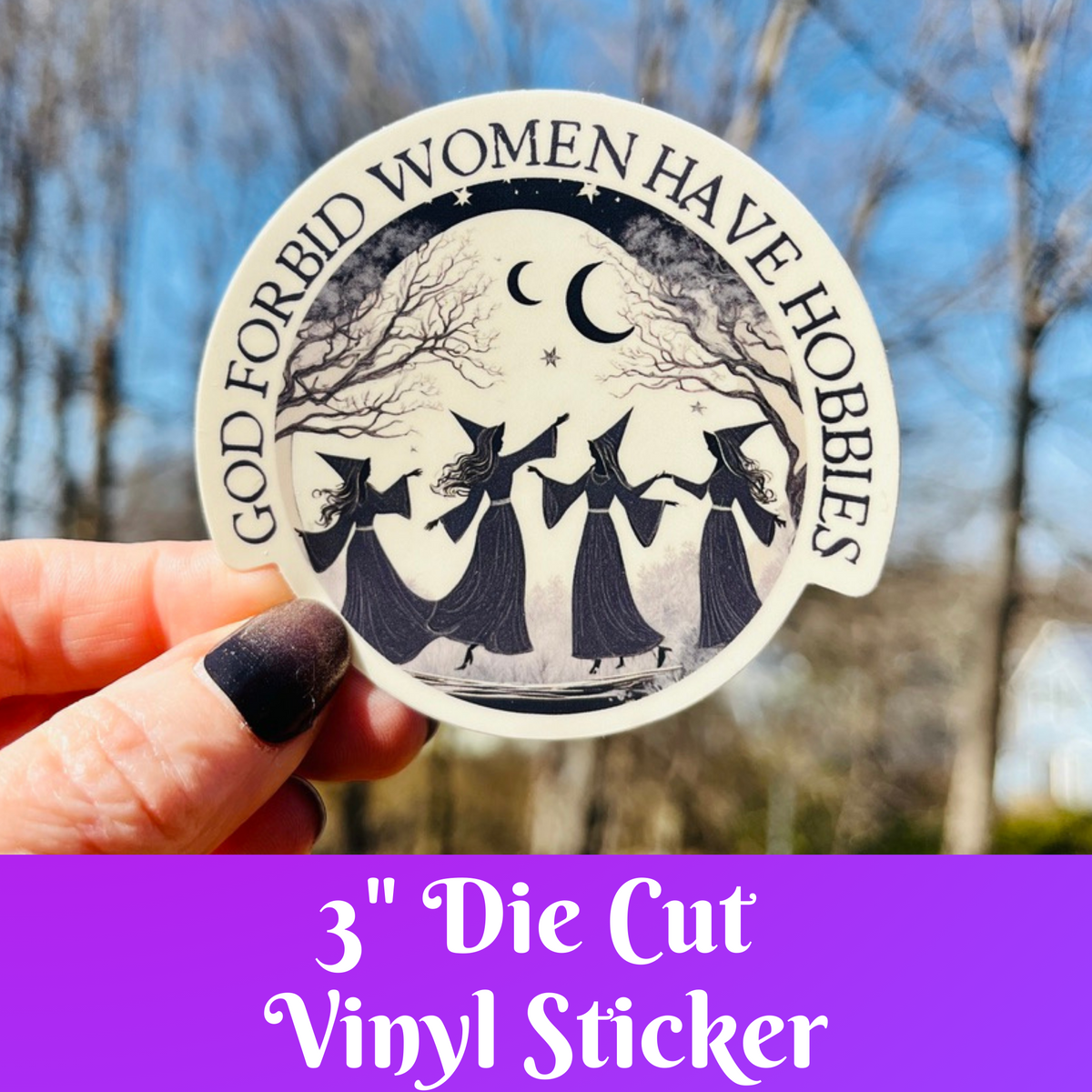 A hand holds a round die-cut vinyl sticker featuring a black and white illustration of three witches dancing under a crescent moon, with the text 'GOD FORBID WOMEN HAVE HORRORS' around the circle. The background is a blurred outdoor scene with bare trees and blue sky, and a purple banner at the bottom reads '3" Die Cut Vinyl Sticker'.