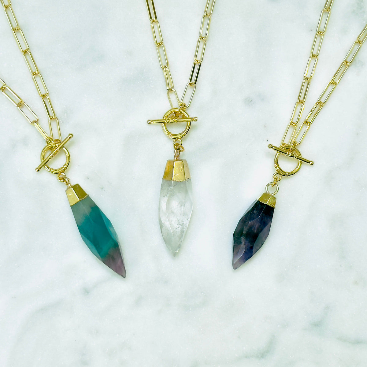 Three gold chain necklaces with toggle clamps and teardrop-shaped crystal pendants are arranged on a white marble surface. The left pendant is teal with a purple gradient at the tip, the center pendant is milky white, and the right pendant is dark blue with a purple gradient at the tip.