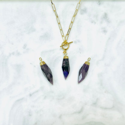 A gold chain necklace with a toggle clasp and three faceted purple crystal pendants in gold caps, displayed on a white marble surface.