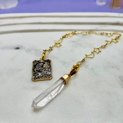 A gold-toned necklace with a rectangular engraved pendant and a clear crystal point pendant, displayed on a white marble surface.