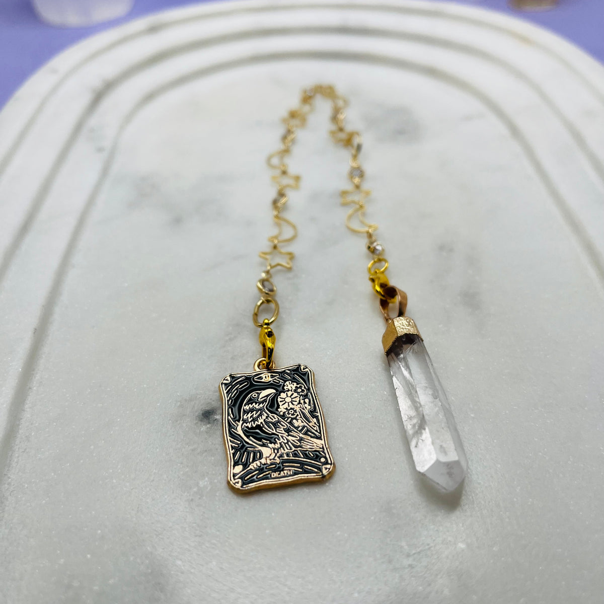 A gold-toned pendant necklace featuring a rectangular engraved charm and a clear crystal point, displayed on a white marble tray.
