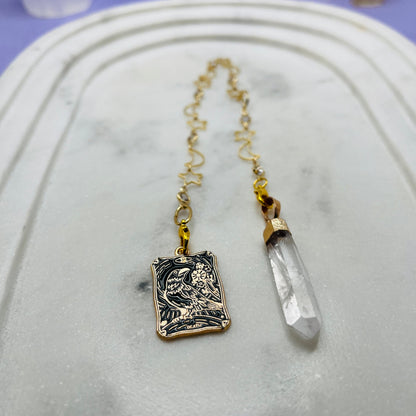 A gold-toned pendant necklace featuring a rectangular engraved charm and a clear crystal point, displayed on a white marble tray.