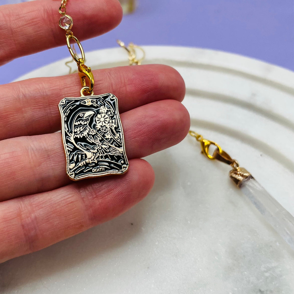 A hand holding a silver-toned rectangular pendant with an engraved owl design, attached to a gold-toned chain with a clear crystal charm, against a white tray and light purple background.