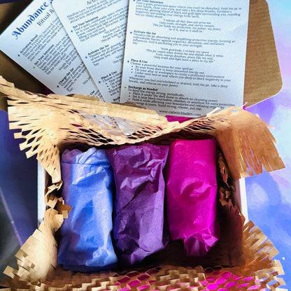 A gift box containing three wrapped spell jars in blue, purple, and pink, with printed cards titled 'Protection Spell Jar', 'Love Spell Jar', and 'Ambition Spell Jar' fanned out above them.