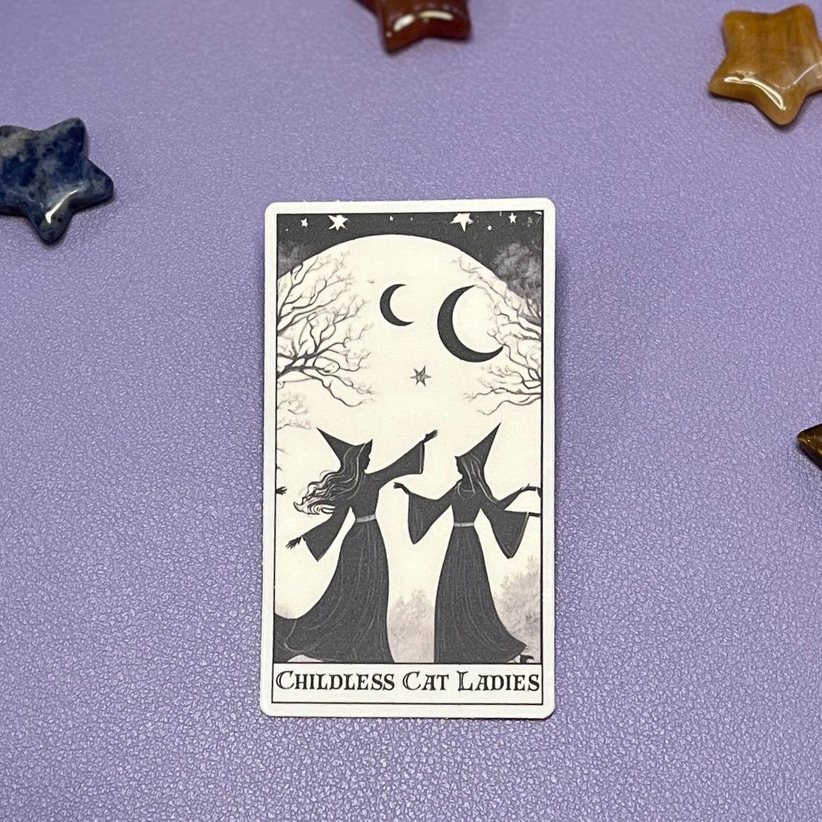A tarot card titled 'Childless Cat Ladies' featuring two witch silhouettes under a crescent moon and stars, displayed on a textured lavender surface with decorative star-shaped stones.