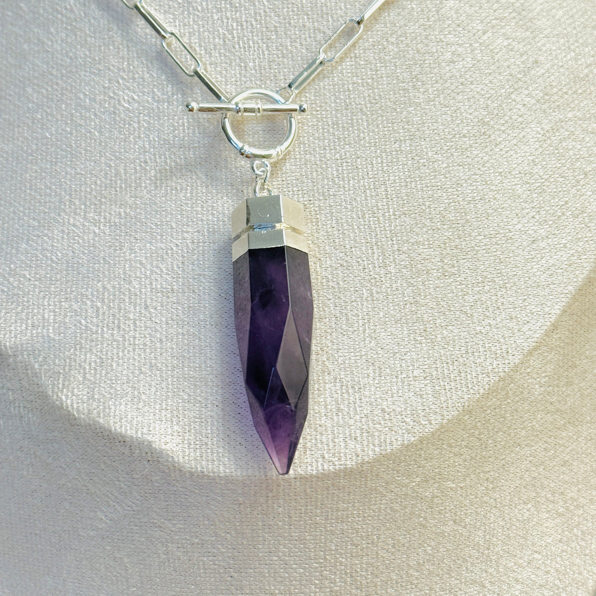 A silver chain necklace with a faceted purple crystal pendant, displayed on a textured cream fabric bust. The pendant is capped with a silver band and suspended from a toggle clasp on a paperclip chain.
