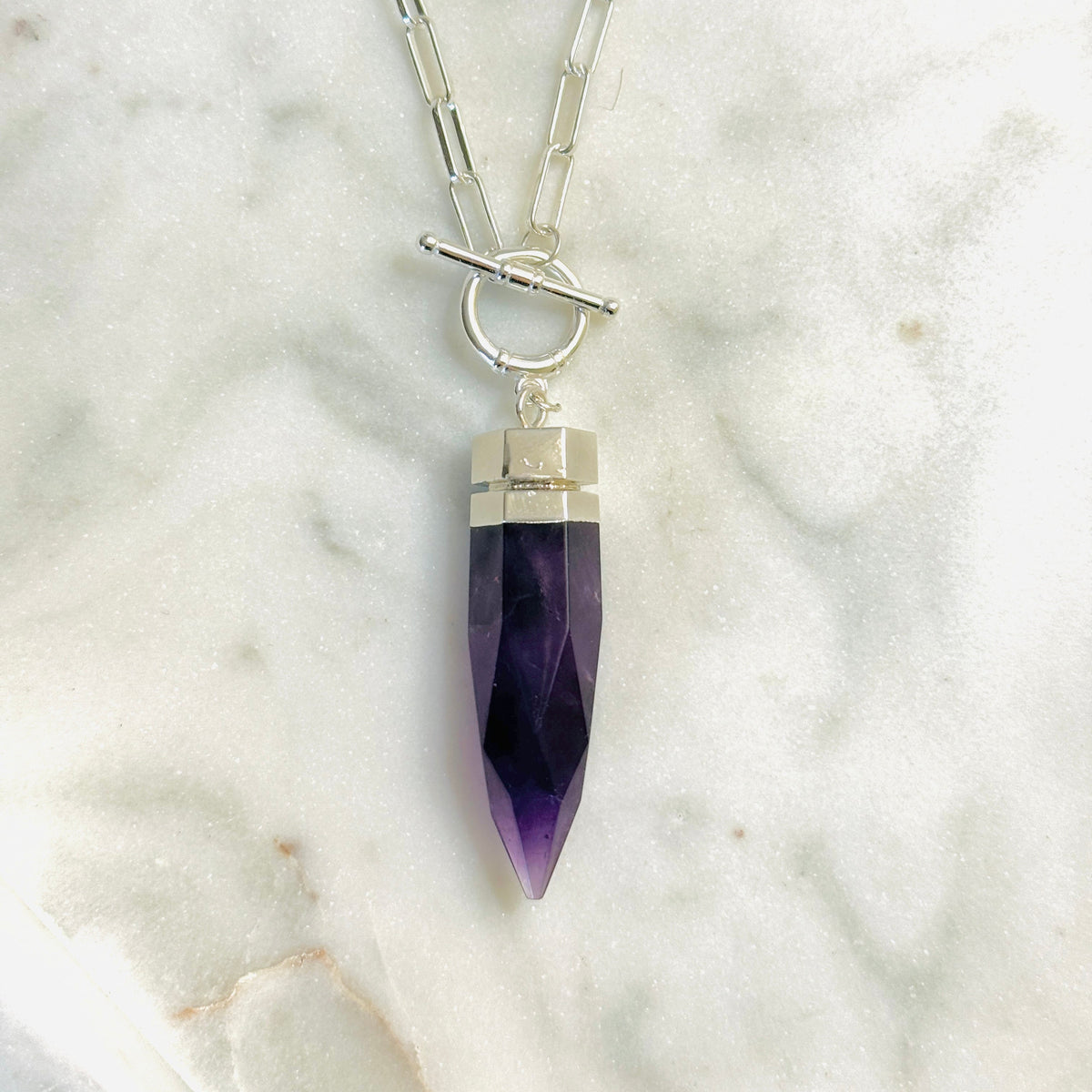 A silver chain necklace with a toggle clasp and a faceted purple crystal pendant, displayed on a white marble surface.