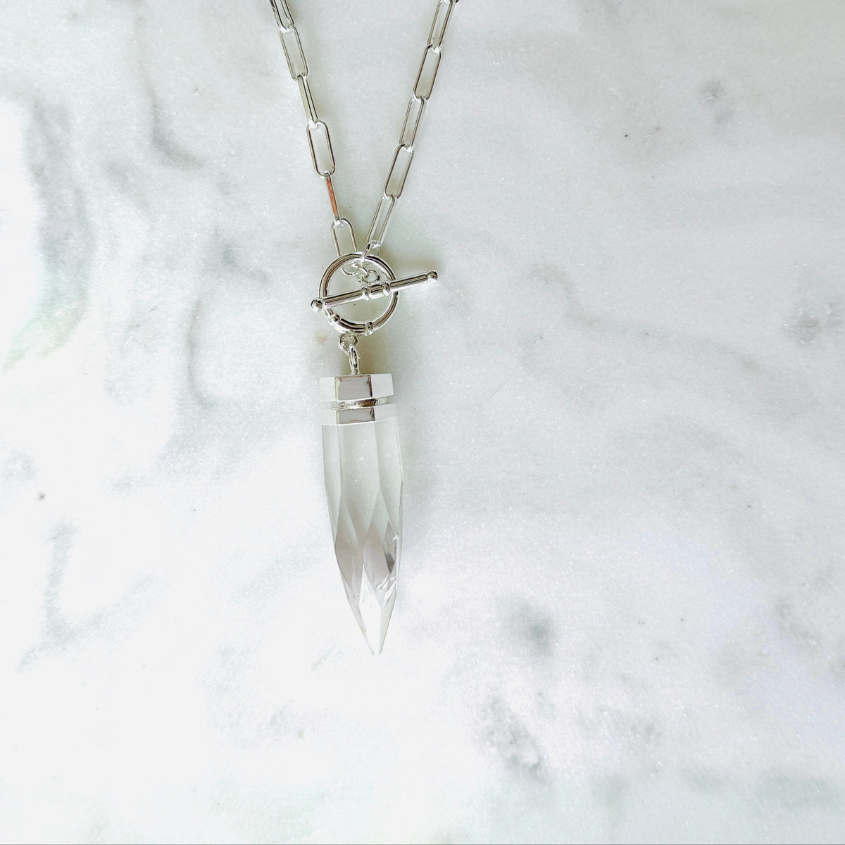 A silver-toned chain necklace with a clear faceted crystal pendant and a toggle clasp, displayed on a light marble surface.