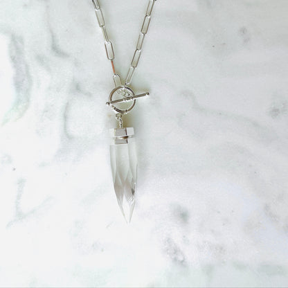 A silver-toned chain necklace with a clear faceted crystal pendant and a toggle clasp, displayed on a light marble surface.