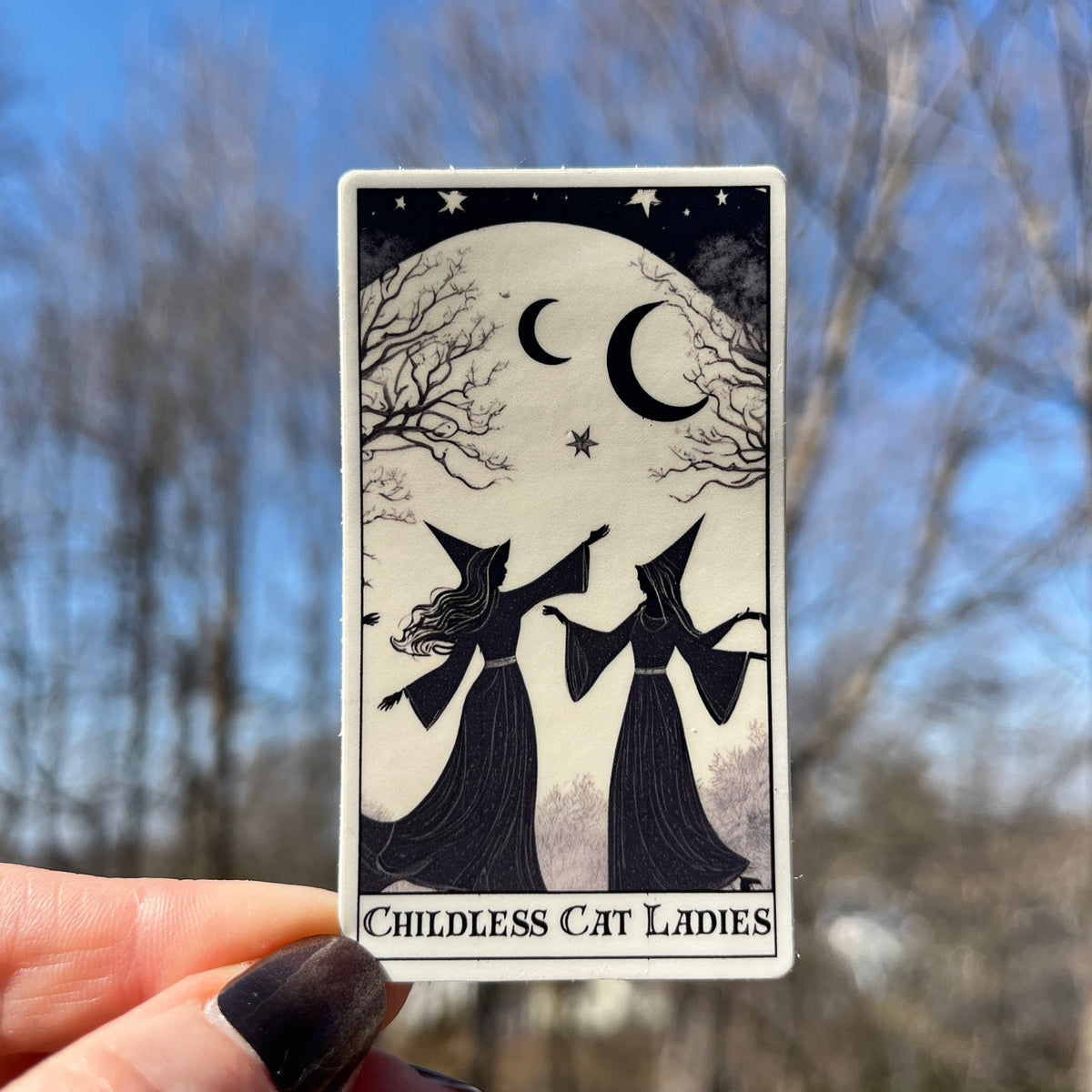 A hand with dark nail polish holds a tarot card titled 'Childless Cat Ladies' against a blurred outdoor background of bare trees and blue sky. The card features a black and white illustration of two witches in pointed hats under a crescent moon and full moon.