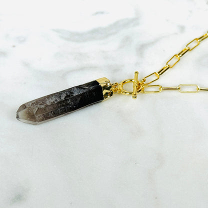 A gold chain necklace featuring a smoky quartz crystal pendant with a gold toggle clasp, displayed on a white marble surface.