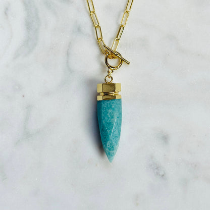 A gold chain necklace with a toggle clasp and a turquoise-colored stone pendant, displayed on a white marble surface.