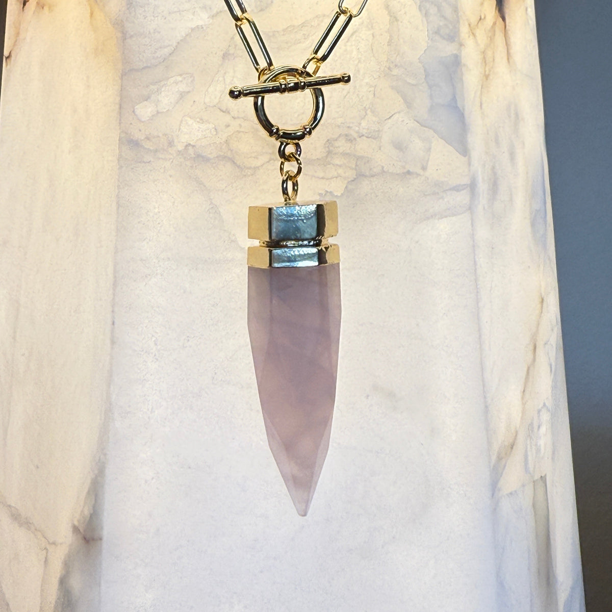 A gold-tone chain necklace with a toggle clasp and a tapered rose quartz crystal pendant, displayed against a white marble column.