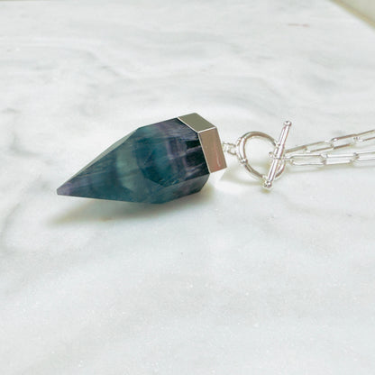 A silver pendant necklace featuring a faceted dark teal-blue gemstone point, resting on a white marble surface with a silver paperclip chain.
