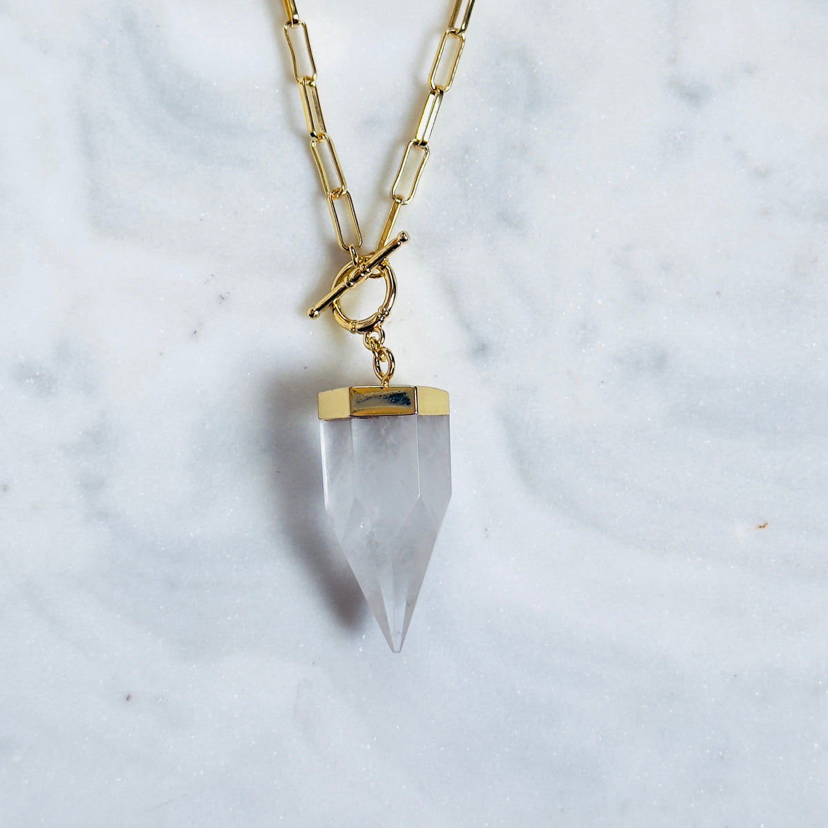 A gold chain necklace with a toggle clasp and a clear pointed crystal pendant, displayed on a white marble surface.