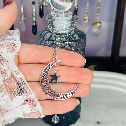 A hand wearing a white lace sleeve holds a silver crescent moon and star pendant in front of a dark glass bottle with a crystal stopper. The pendant features an ornate filigree crescent moon with a small dangling star charm.