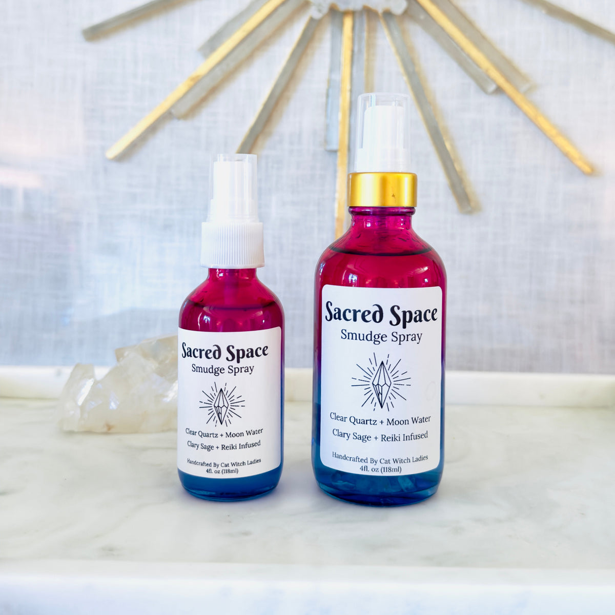 Two bottles of Sacred Space Smudge Spray displayed on a white marble surface with a white textured wall and a gold sunburst mirror in the background. One bottle is filled with blue liquid and the other with pink liquid, both with white labels and white spray tops.