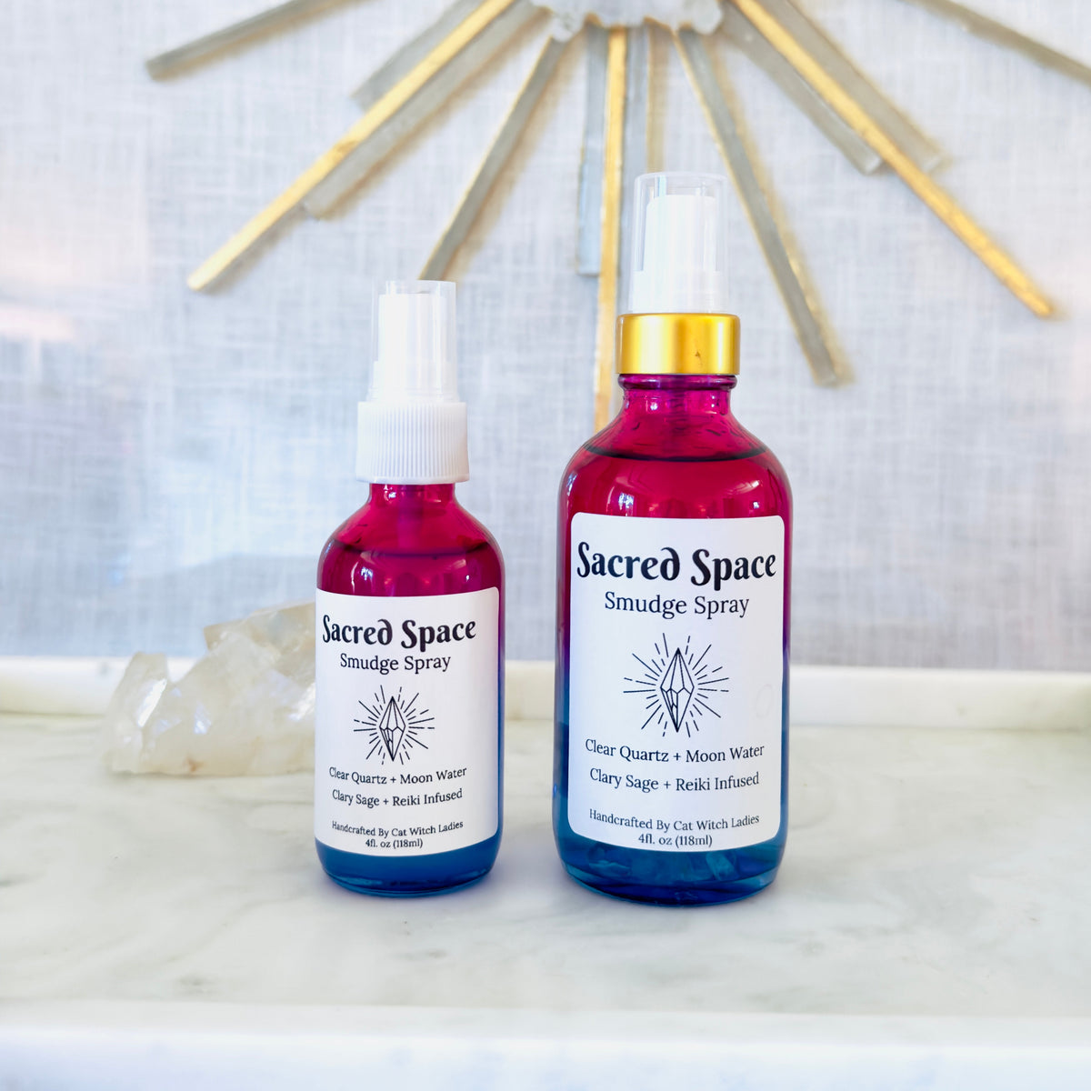 Two bottles of Sacred Space Smudge Spray displayed on a white marble surface with a white and gold sunburst wall mirror in the background.