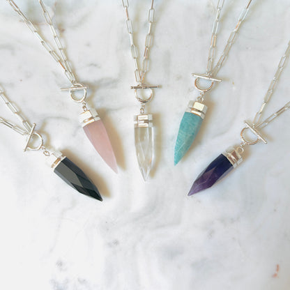 Four silver-toned spike pendant necklaces with different colored crystal points are arranged on a white marble surface. The necklaces feature black, pink, clear, and teal crystals.