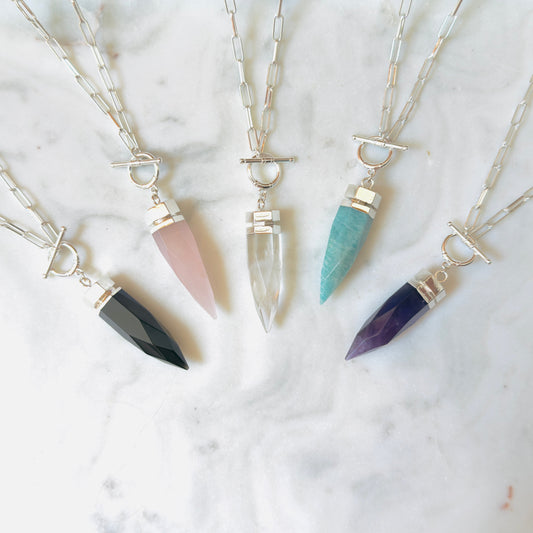 Four silver-toned spike pendant necklaces with different colored crystal points are arranged on a white marble surface. The necklaces feature black, pink, clear, and teal crystals.