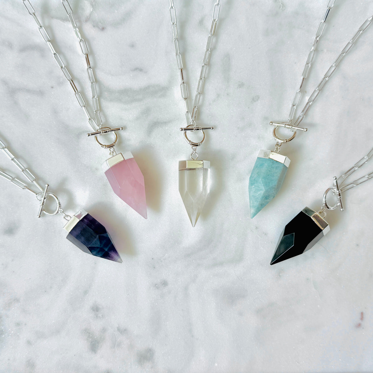 Four crystal point pendant necklaces with silver chains and toggle closures, arranged on a white marble surface. The crystals are colored dark blue, pink, clear, and black.