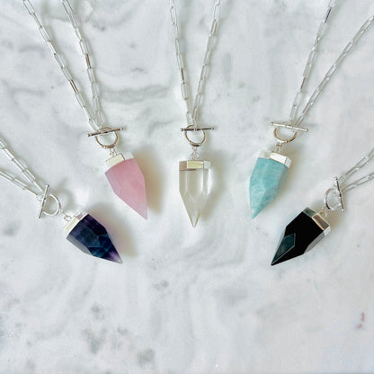 Four crystal point pendant necklaces with silver chains and toggle closures, arranged on a white marble surface. The crystals are colored dark blue, pink, clear, and black.