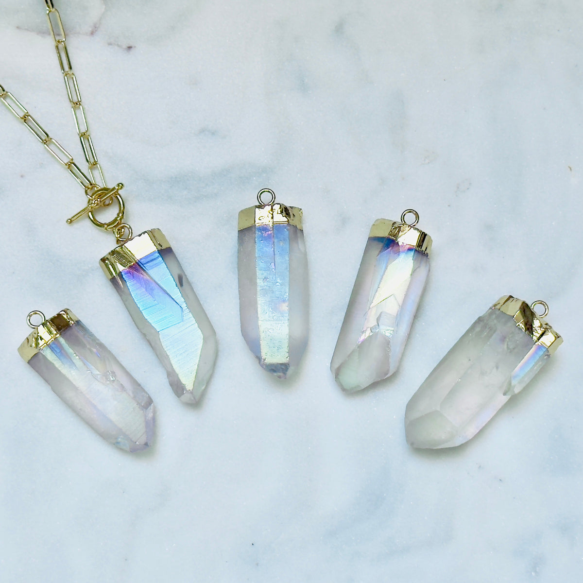 Five iridescent crystal point pendants with gold caps are arranged on a white marble surface, with a gold chain and clasp visible in the upper left.