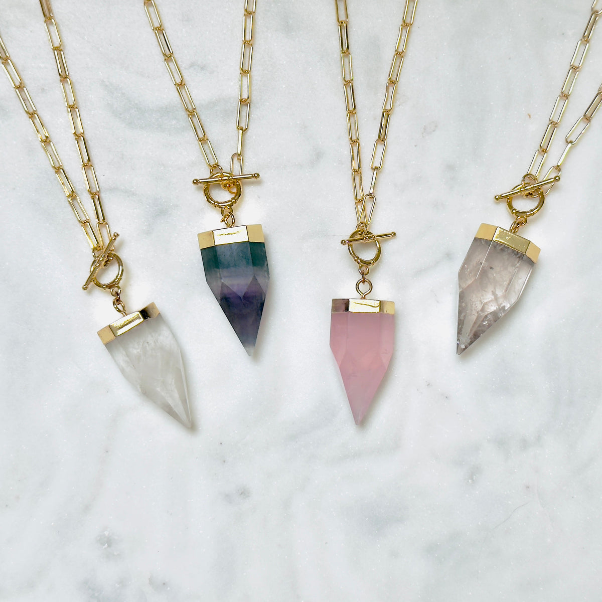 Four gold chain necklaces with toggle clasp and pointed crystal pendants in white, green-to-purple, pink, and smoky gray, arranged on a white marble surface.