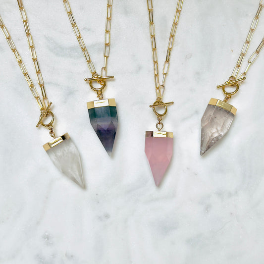 Four gold chain necklaces with toggle clasp and pointed crystal pendants in white, green-to-purple, pink, and smoky gray, arranged on a white marble surface.