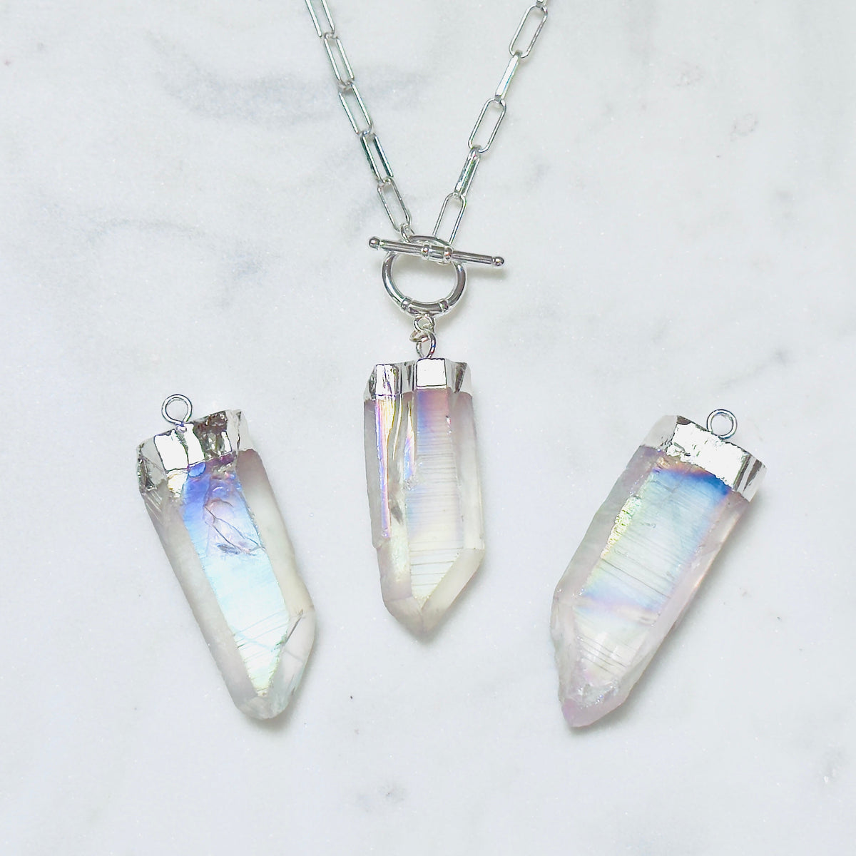 Three silver-toned crystal point pendants on a silver chain, displayed on a white marble surface. The crystals have an iridescent, milky white appearance with subtle pastel rainbow reflections.