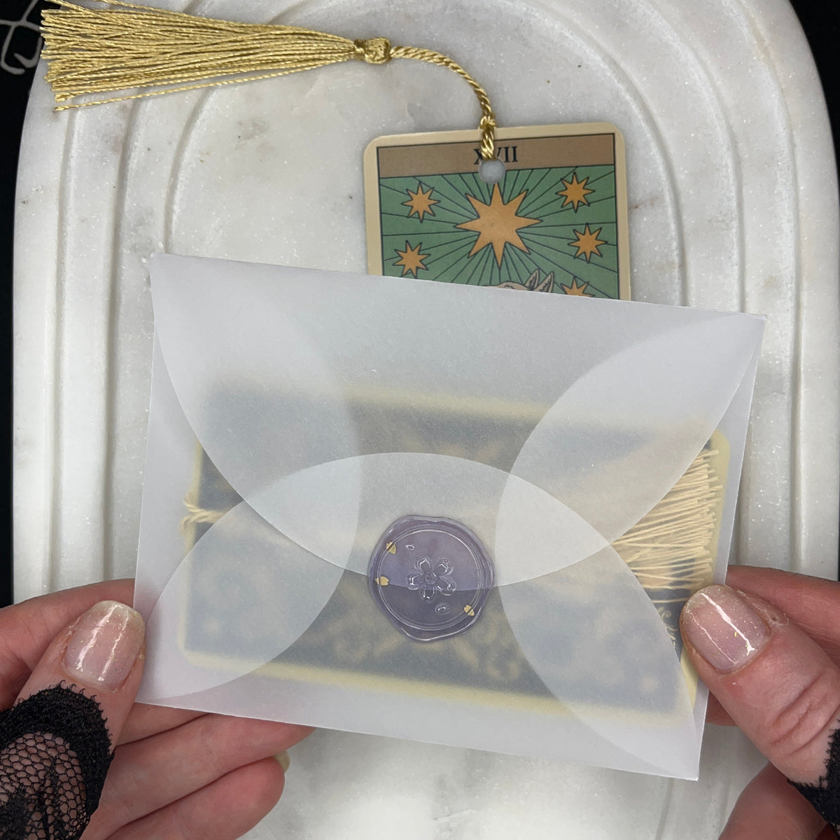 A person's hands hold a translucent white envelope with a purple wax seal, containing a tarot card. The card is labeled 'VII' and features a gold star design on a green background, complete with a gold tassel.
