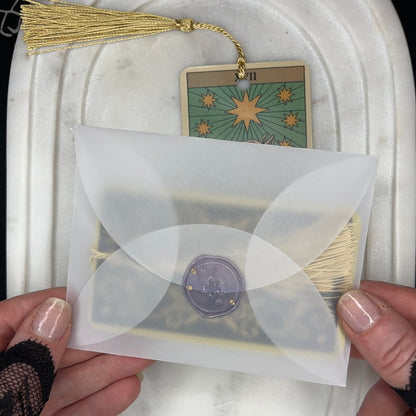 A person's hands hold a translucent white envelope with a purple wax seal, containing a tarot card. The card is labeled 'VII' and features a gold star design on a green background, complete with a gold tassel.