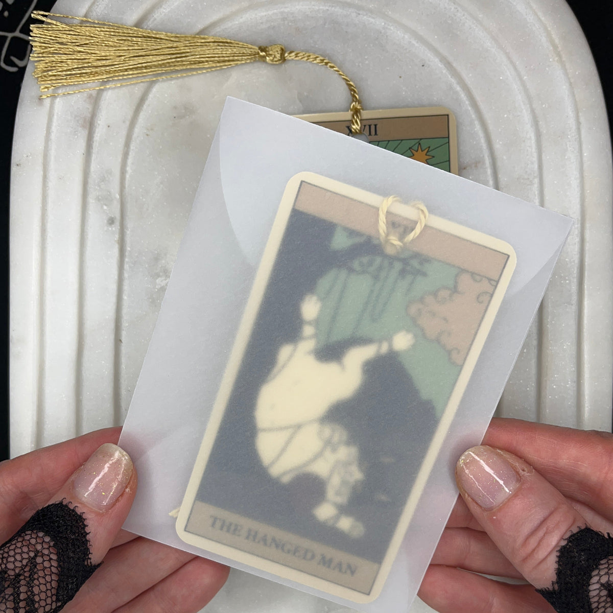 A person holds a tarot card deck in a translucent sleeve, showing the 'The Ranged Man' card. The card has a gold tassel and is displayed over a white marble tray.