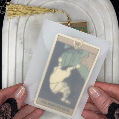 A person holds a tarot card deck in a translucent sleeve, showing the 'The Ranged Man' card. The card has a gold tassel and is displayed over a white marble tray.