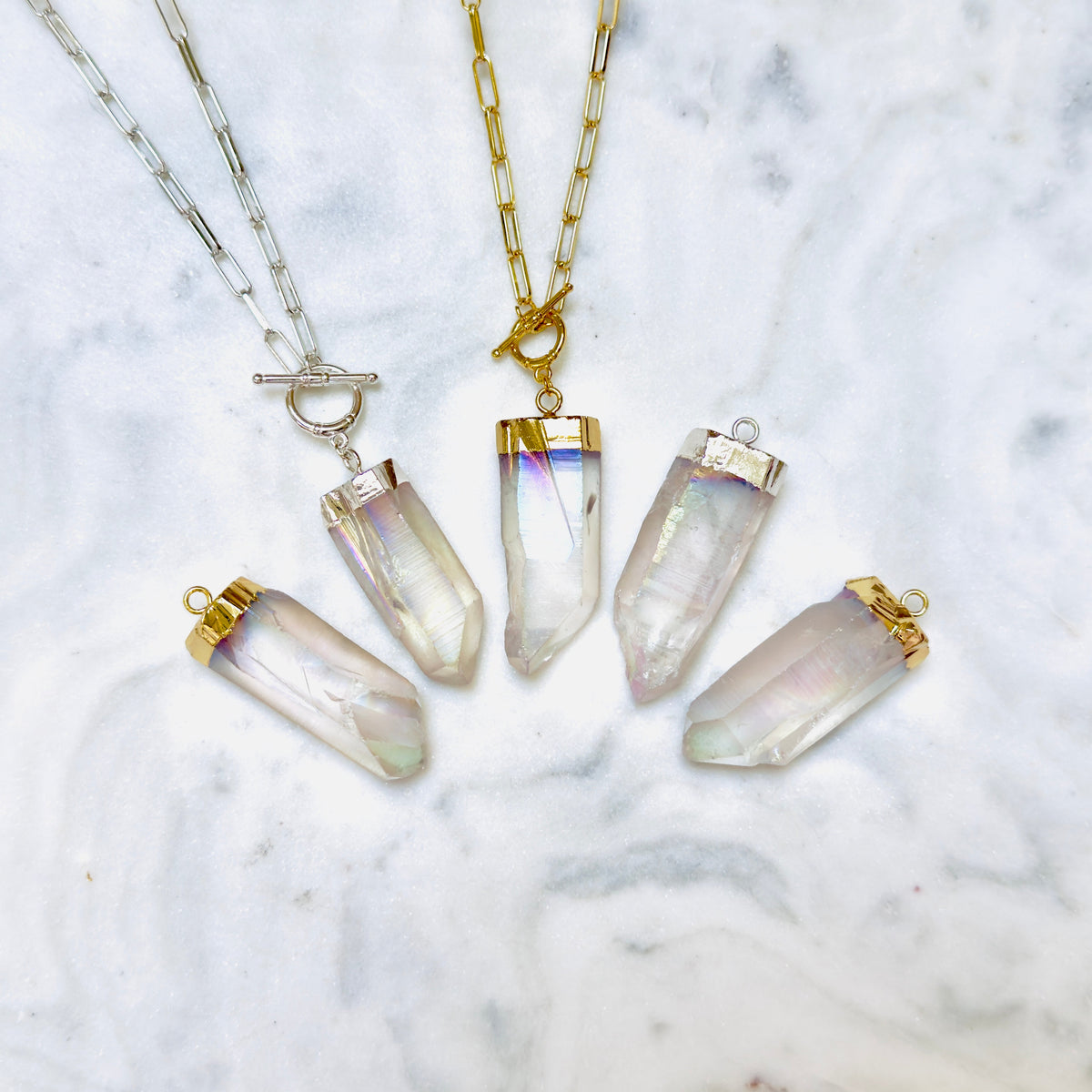Four crystal point pendant necklaces arranged on a white marble surface. Two necklaces have gold-tone chains and caps with clear iridescent crystals, and two have silver-tone chains and caps with clear iridescent crystals.