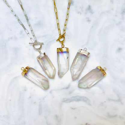Four crystal point pendant necklaces arranged on a white marble surface. Two necklaces have gold-tone chains and caps with clear iridescent crystals, and two have silver-tone chains and caps with clear iridescent crystals.