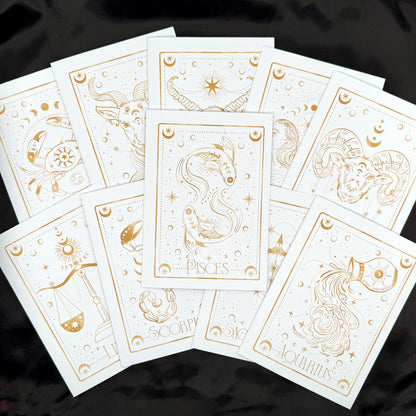 A fan arrangement of white zodiac tarot cards with elegant gold line art and titles including Pisces, Scorpio, Aquarius, and others, displayed on a black fabric background.