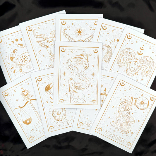 A fan arrangement of white zodiac tarot cards with elegant gold line art and titles including Pisces, Scorpio, Aquarius, and others, displayed on a black fabric background.