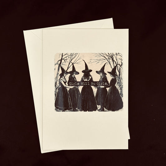 A cream-colored greeting card with a black vintage-style illustration of five witches holding a banner that reads 'Girls Will Be Girls.' The card is shown with its matching envelope against a dark brown background.