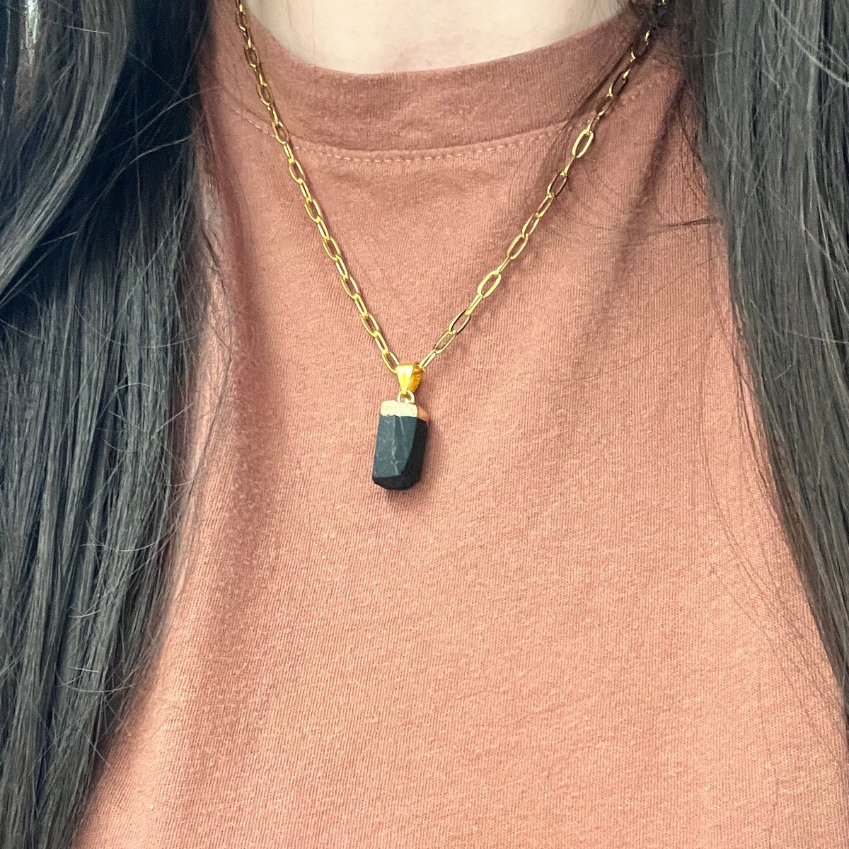 A close-up of a person wearing a gold chain necklace with a dark green faceted rectangular pendant, styled over a dusty rose top.