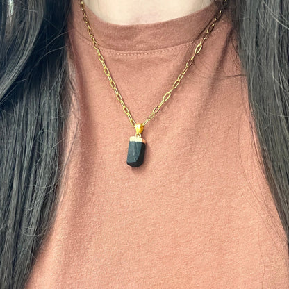 A close-up of a person wearing a gold chain necklace with a dark green faceted rectangular pendant, styled over a dusty rose top.