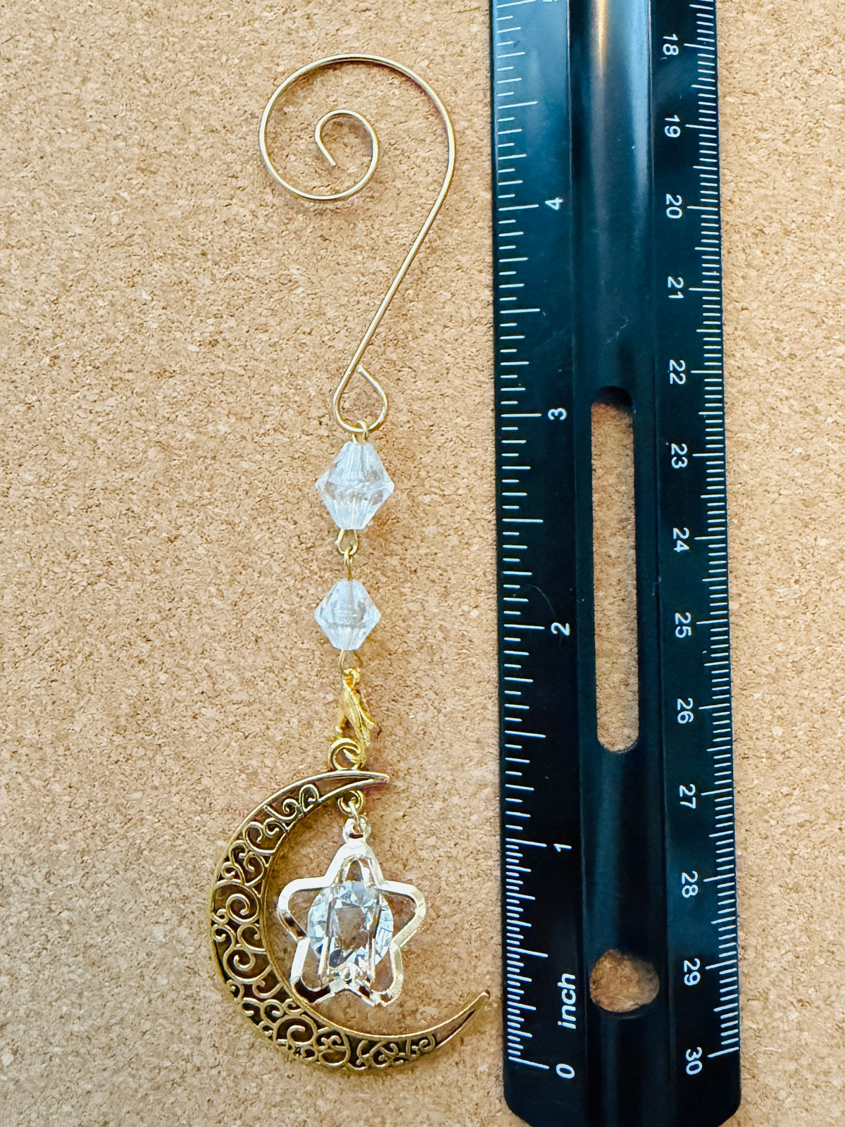 A gold-toned metal bookmark or hair hook with a spiral top, two clear faceted beads, and a dangling crescent moon and star charm, shown next to a black ruler on a tan cork-like background.