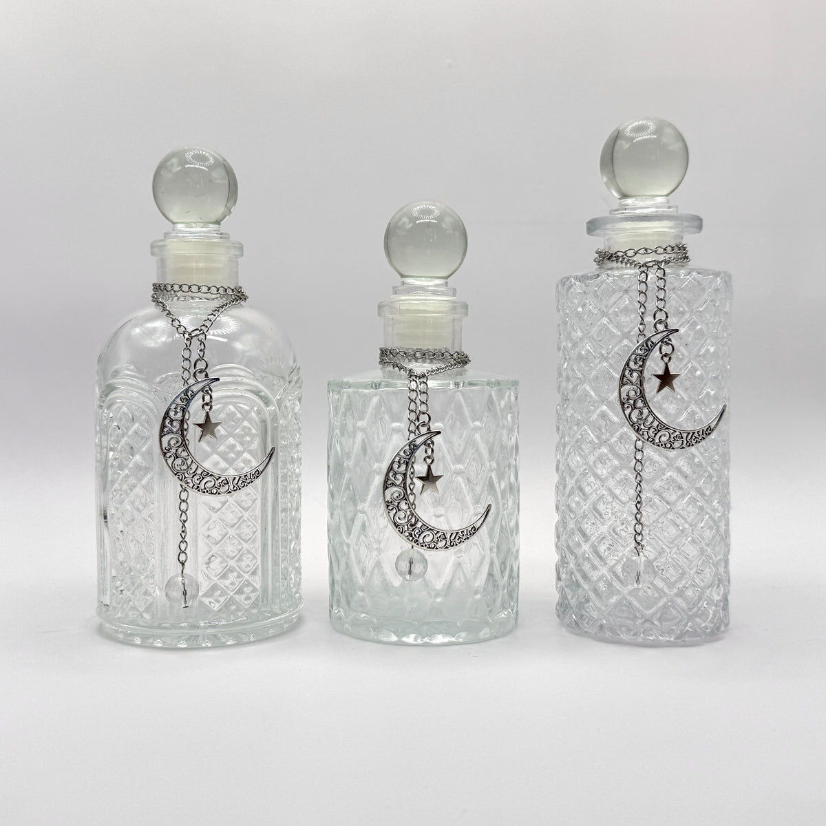 Three clear glass perfume bottles with stoppers and silver crescent moon and star chain ornaments, displayed against a light gray background.