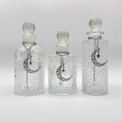 Three clear glass perfume bottles with stoppers and silver crescent moon and star chain ornaments, displayed against a light gray background.